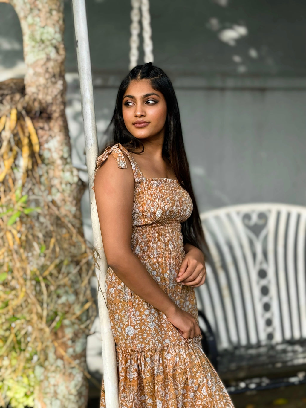 Woman in Hoya Tie-Straps Sun Dress standing outdoors near a tree and chair.